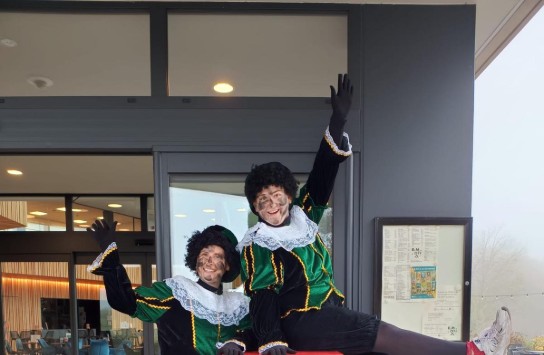 Twee mensen in Pietenkostuum poseren vrolijk bij een rode Hemelbus brievenbus voor Sinterklaas. (vergroot foto)