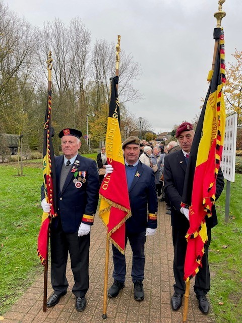Drie mannen in uniformen met vlaggen, staand op een pad tijdens een herdenkingsbijeenkomst. (vergroot foto)